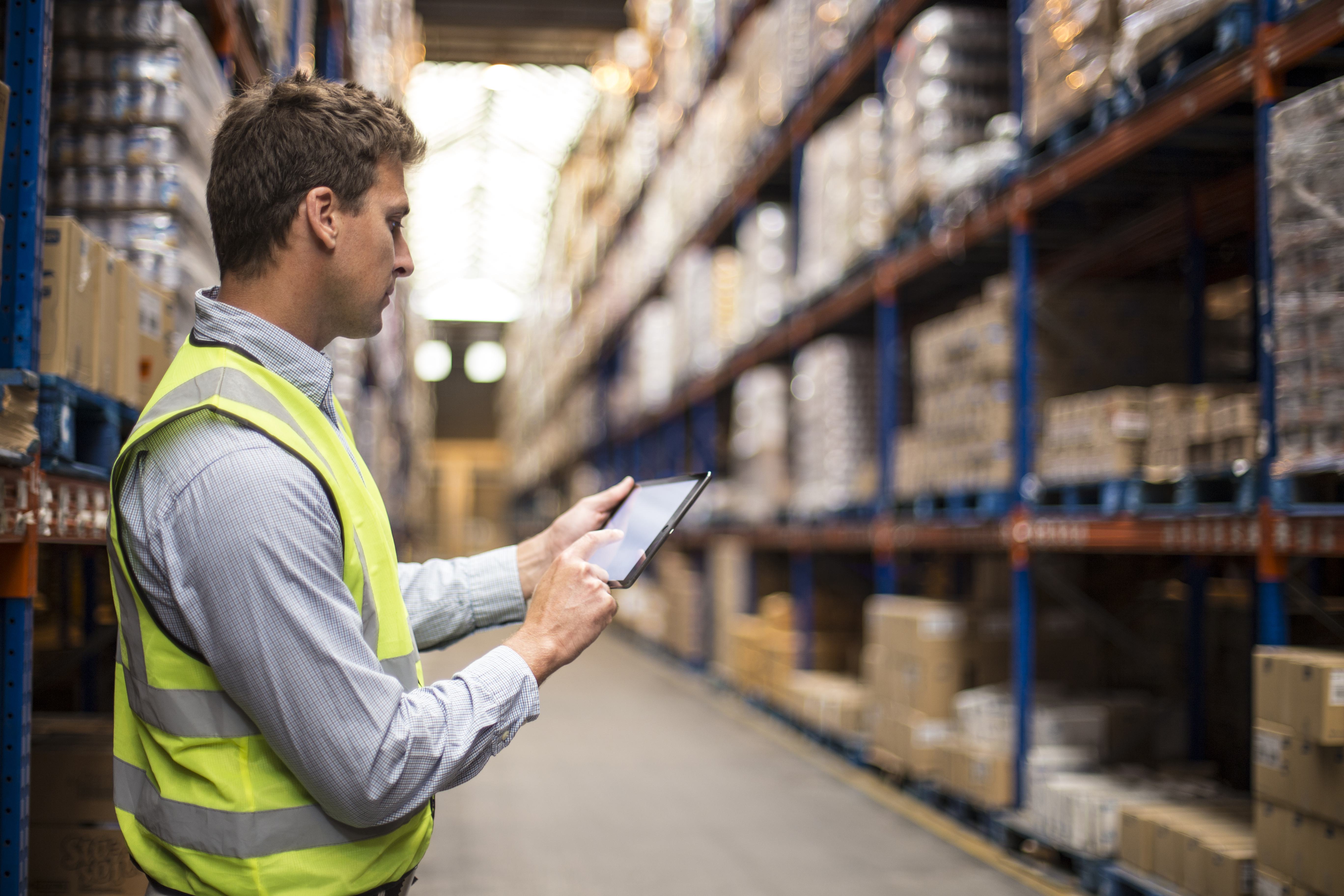 A man wearing a yellow vest stands in a warehouse with shelves full of boxes and looks at a tablet.