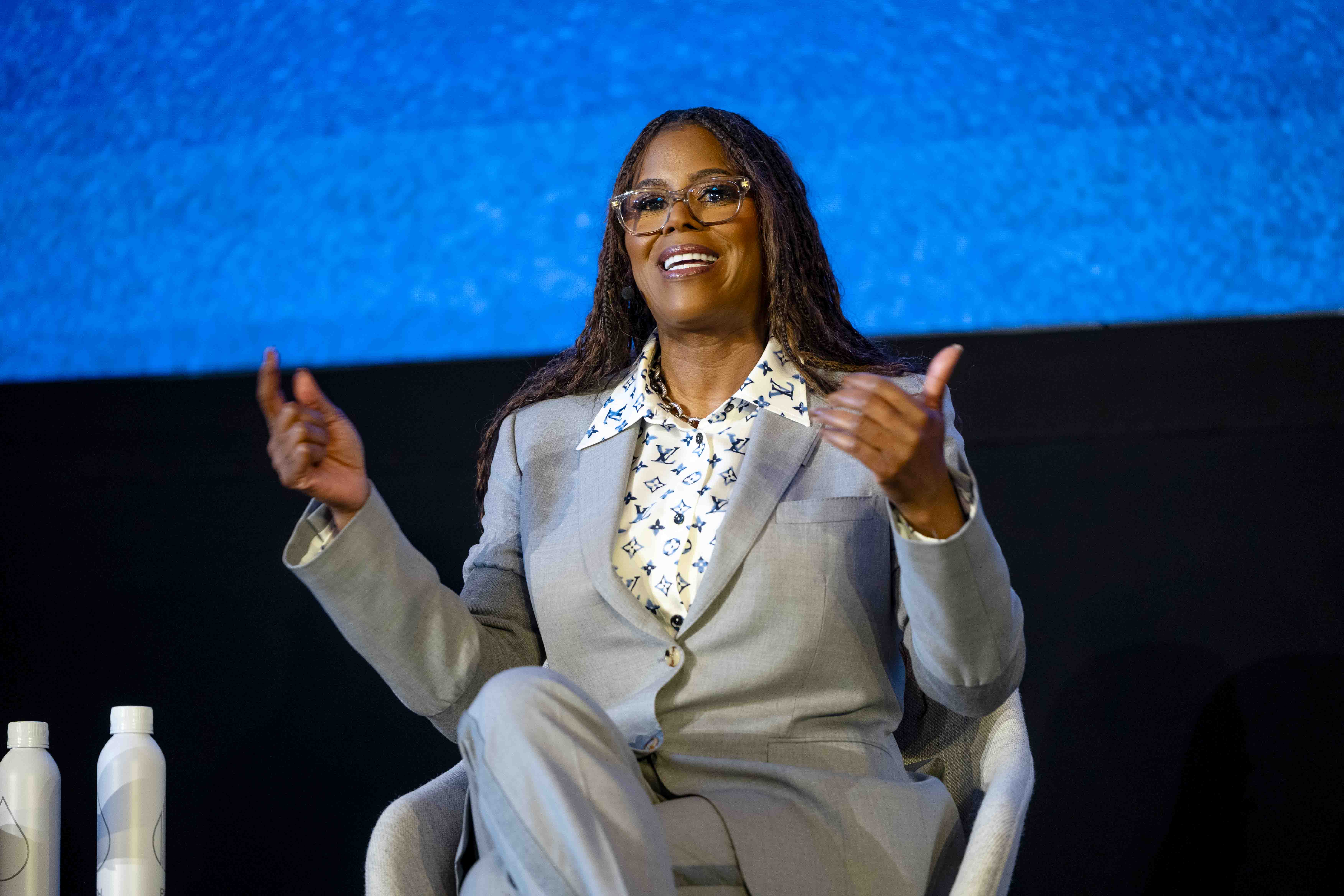 A woman in a gray suit is on-stage during a speech, smiling and gesturing with her hands