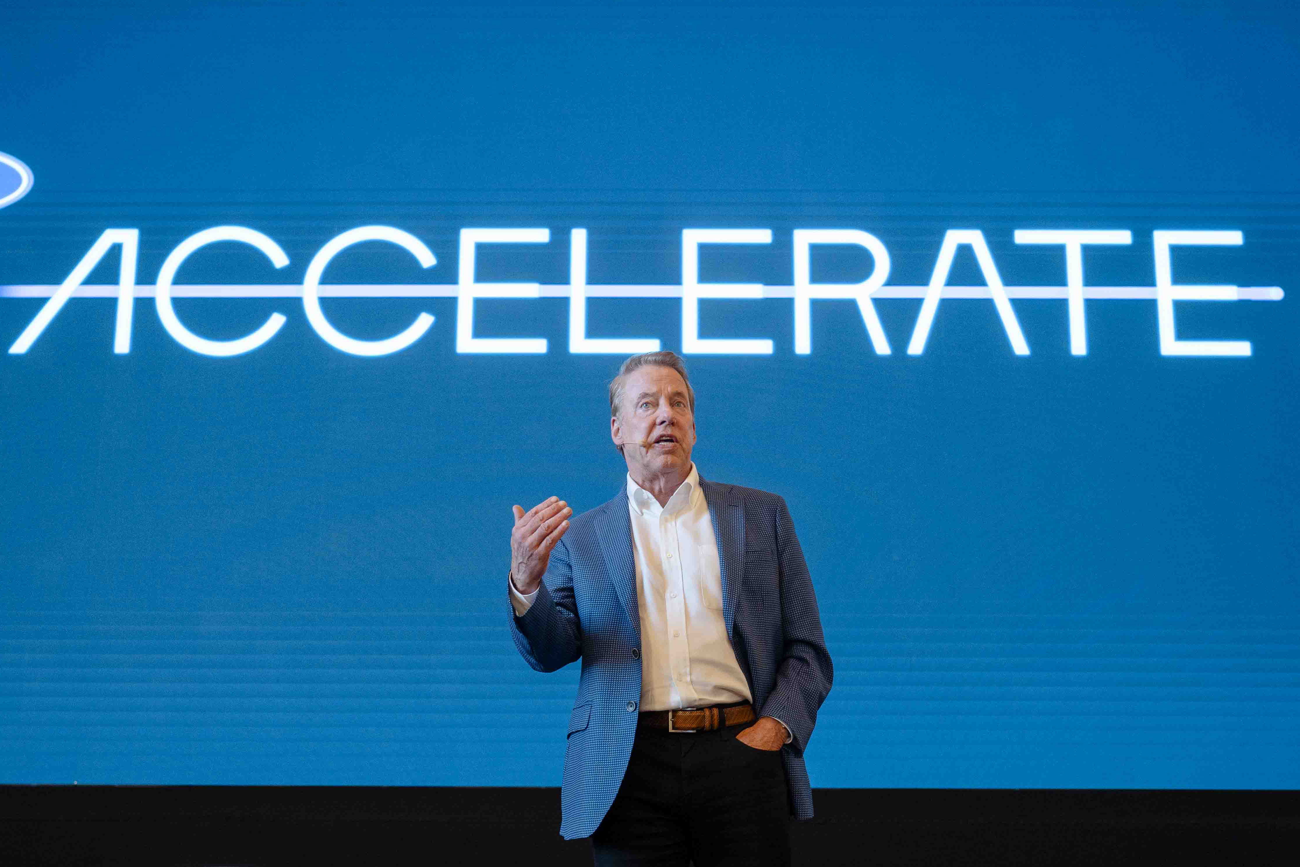 A man in business attire stands on a stage with a backdrop behind him that says "ACCELERATE"