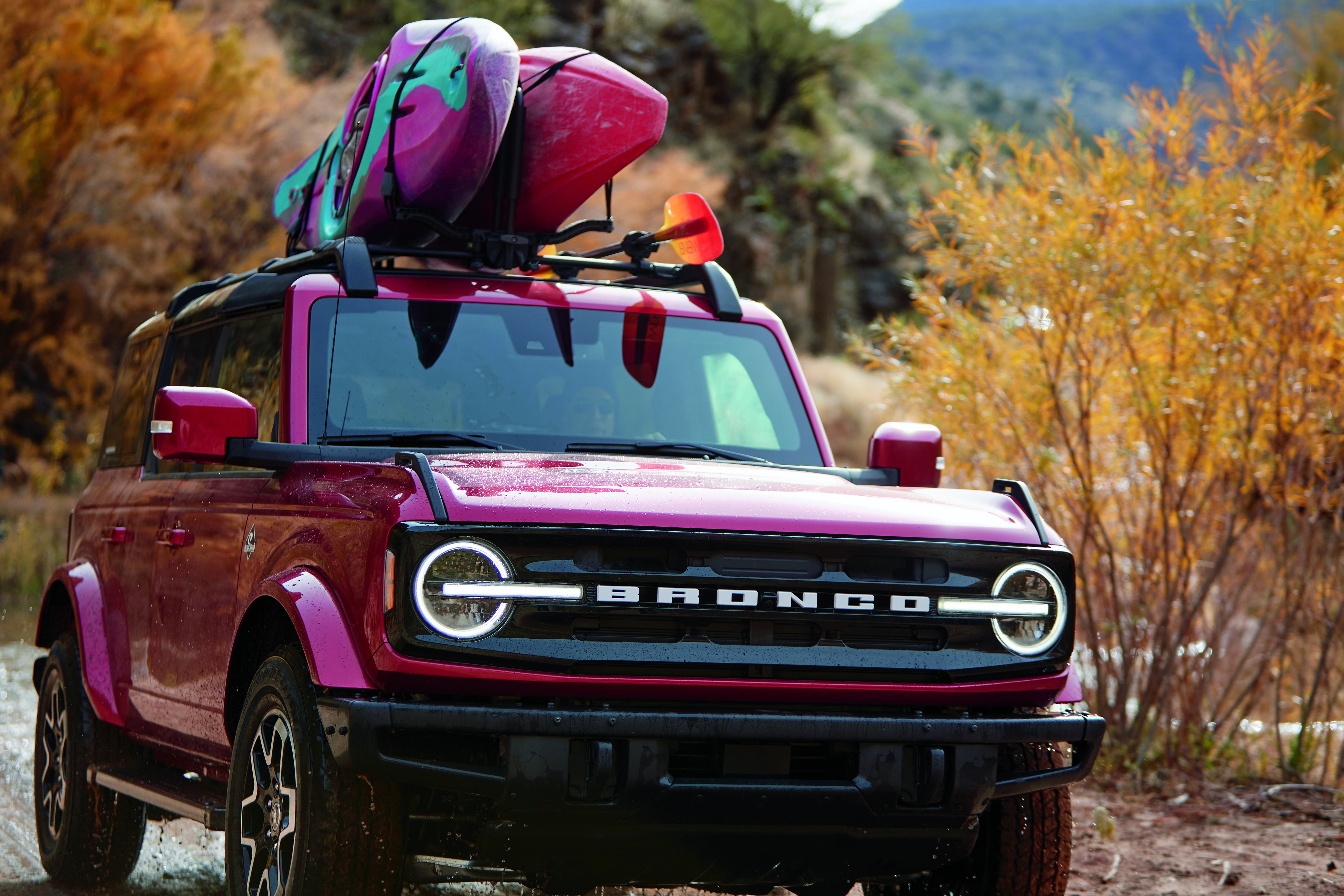 A pinkish purple Ford Bronco drives through a wooded area with kayaks on top if it.