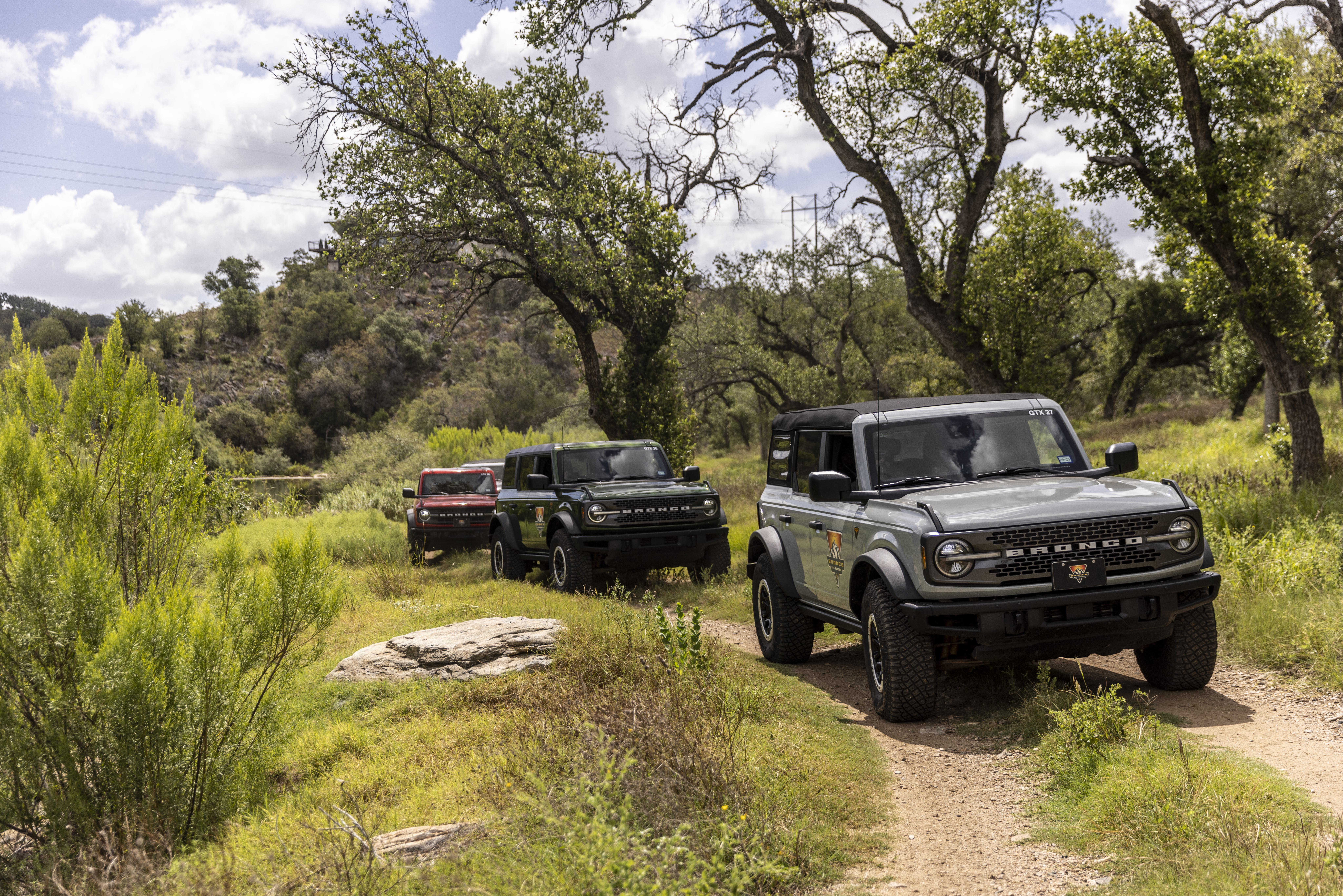 Three Ford Broncos drive on a dirt path through a green, wooded area