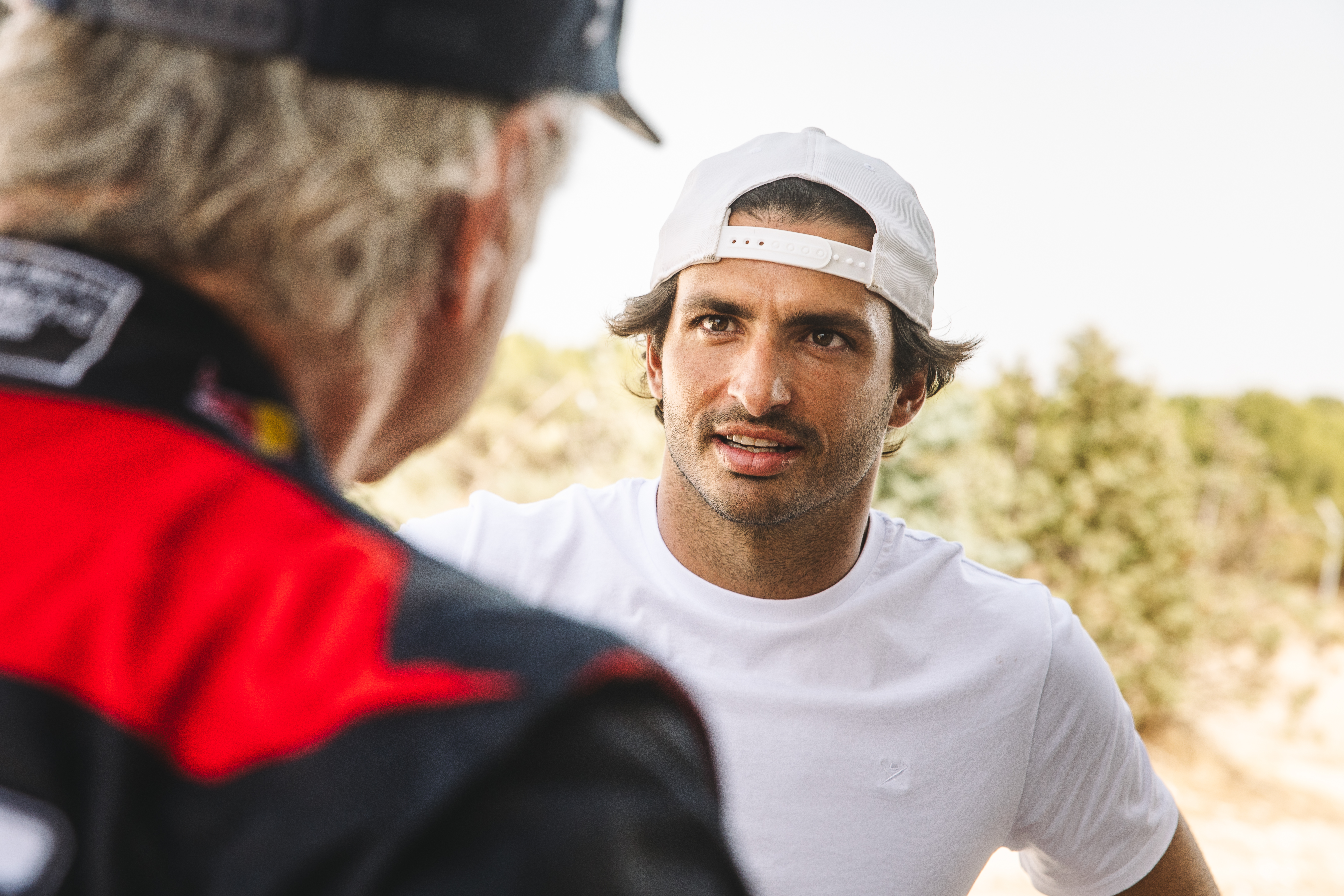 Carlos Sainz is looking toward the camera over the shoulder of his father, Carlos Sainz Senior, who has his back to the camera