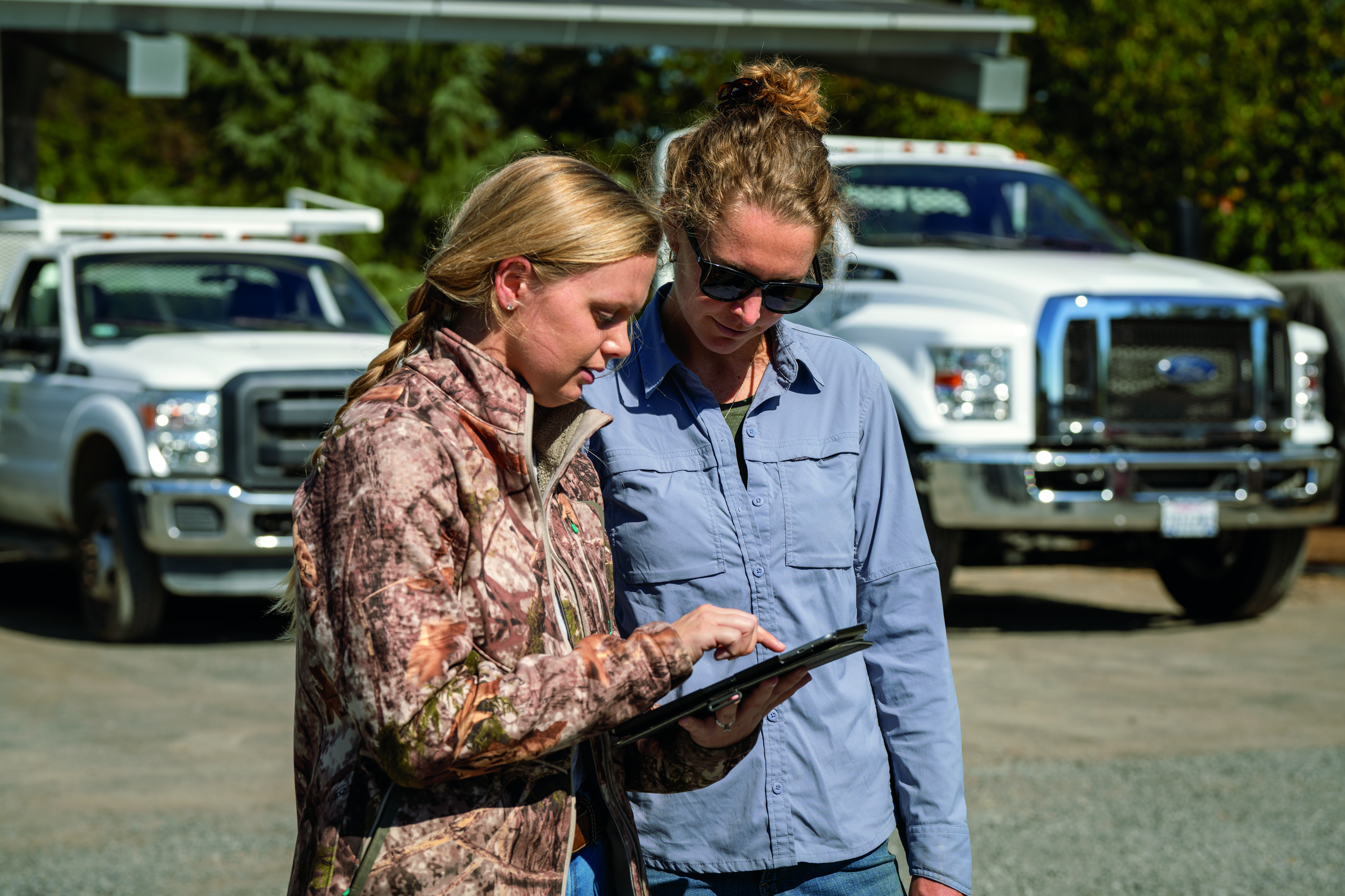 Two women look at a tablet screen in a parking lot. They stand in front of two white trucks.