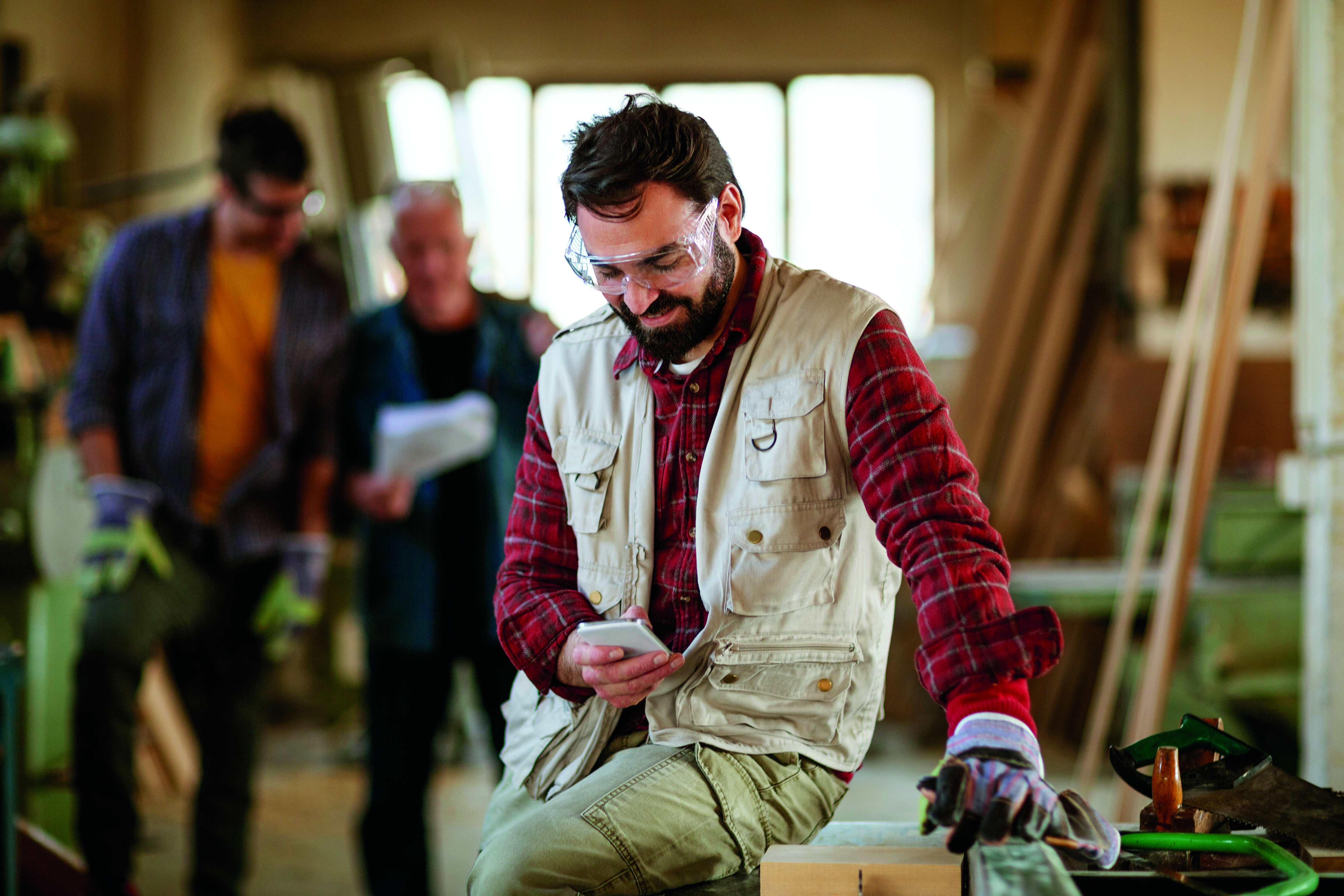 A man wearing protective goggles and a workvest in a workshop looks at his phone
