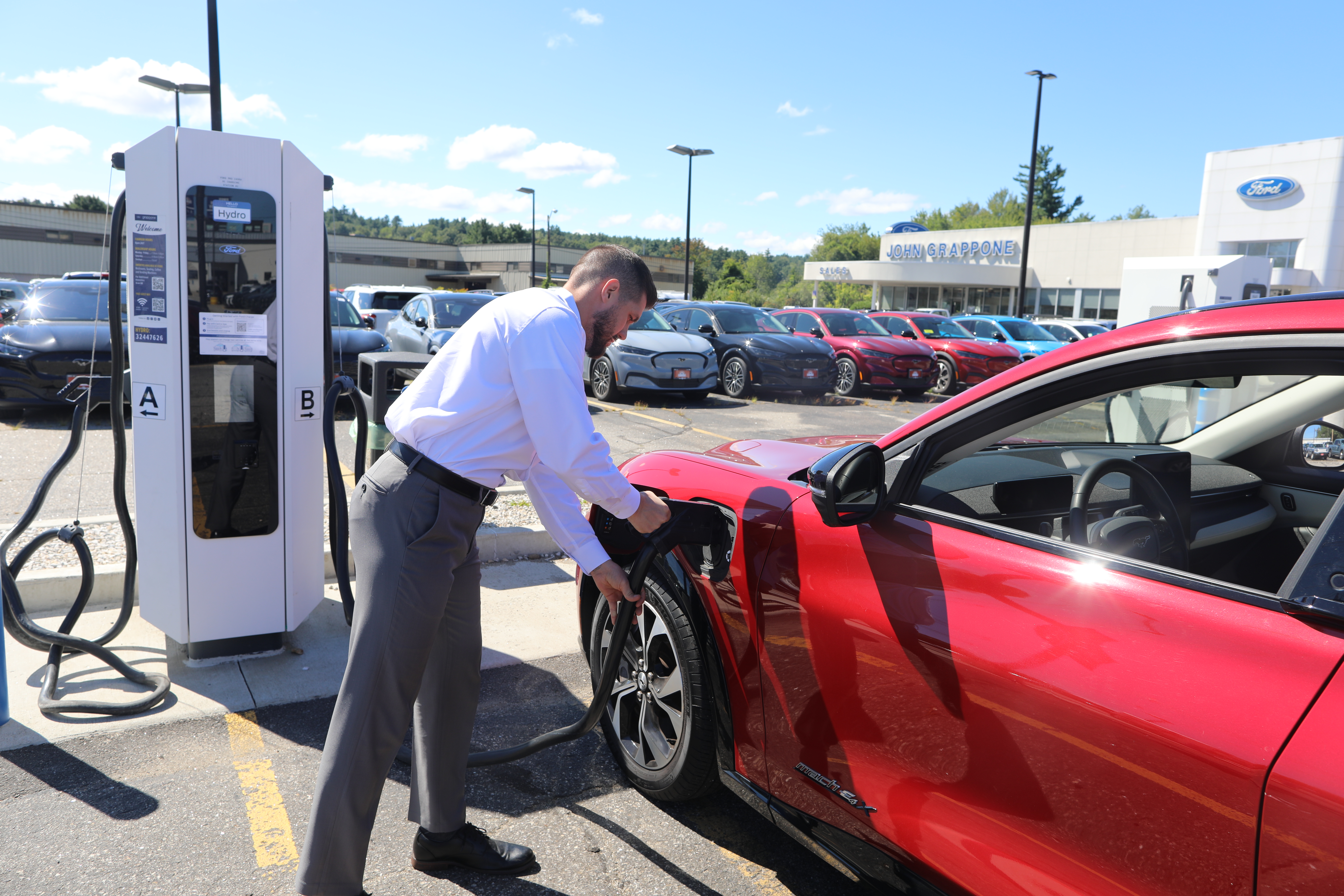 A man charges his red vehicle at an electric vehicle charger in a Ford dealership parking lot.