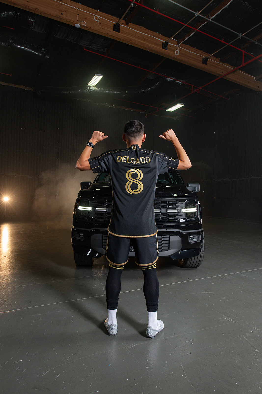 Los Angeles Football Club soccer player Mark Delgado poses in front of the Ford F-150 Lobo truck with his back facing the camera, showing his last name, "Delgado," displayed on his jersey.