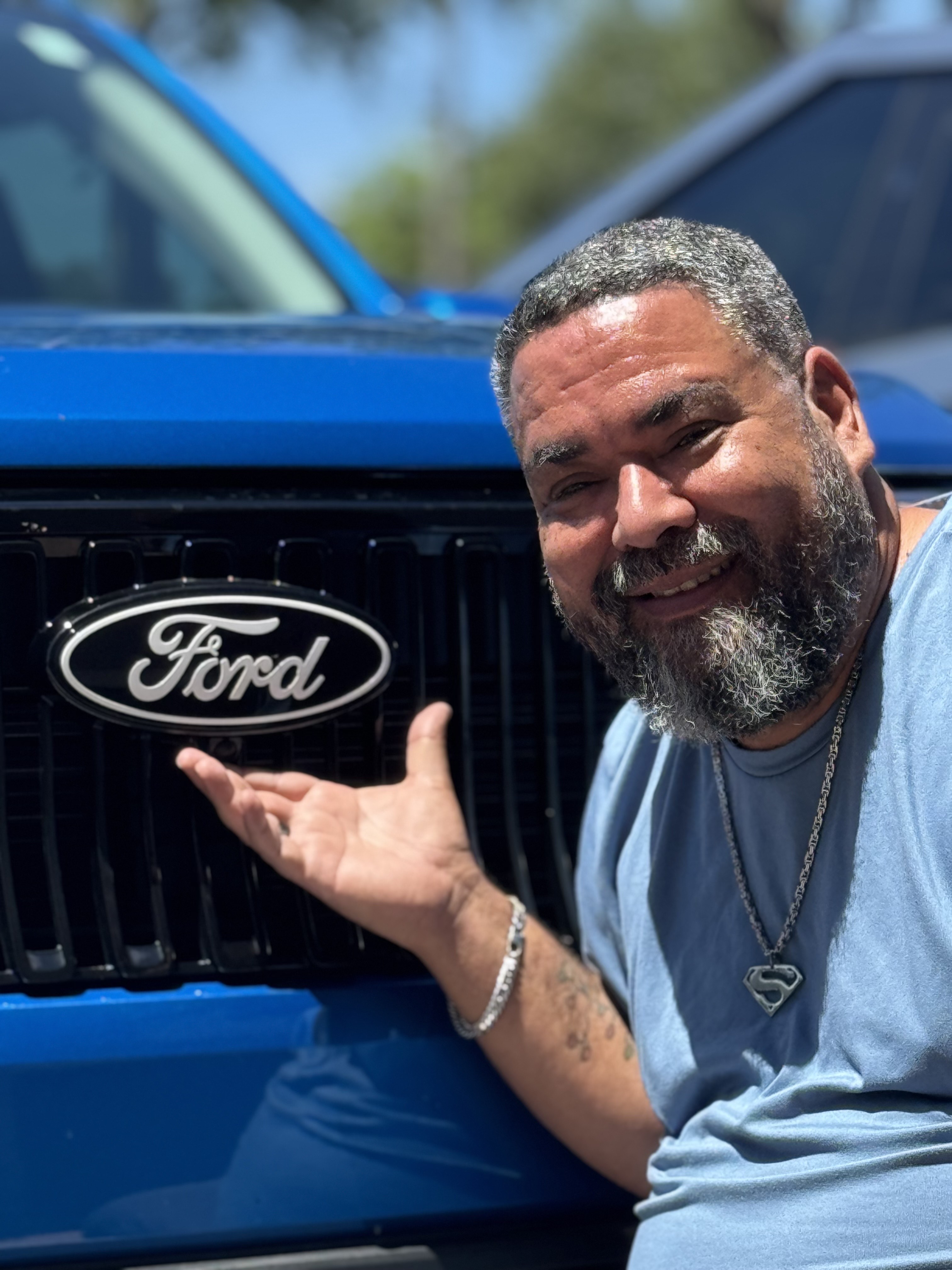 A man poses, smiling, with his blue Maverick Lobo. His hand is open beneath the Ford logo on the front grille.