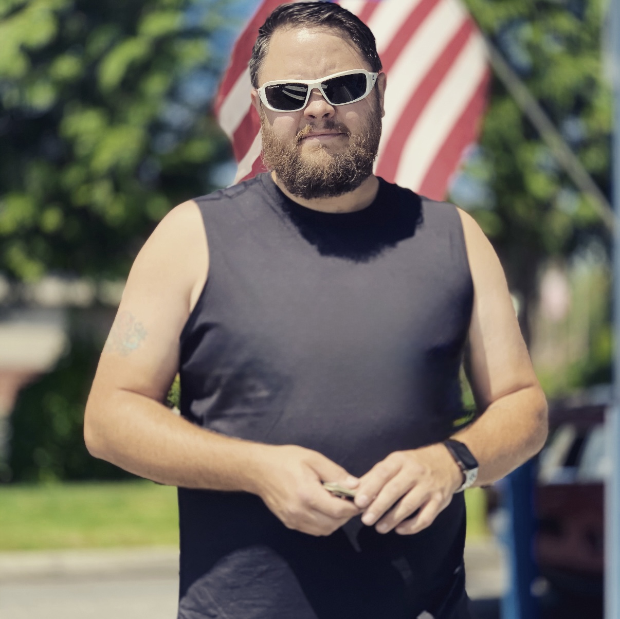 A man in sunglasses and a muscle tee poses in front of an American flag.