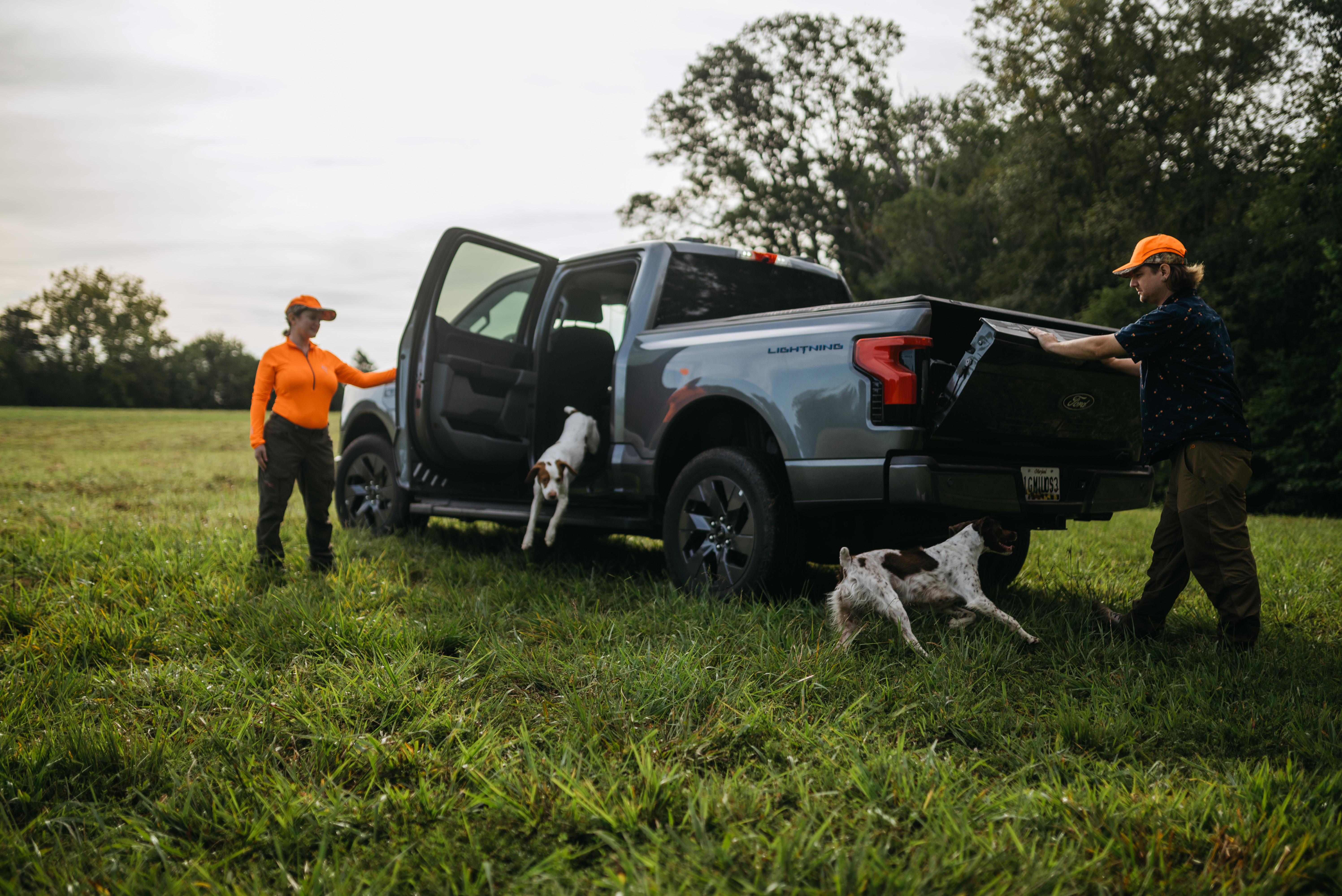 Two hunters, a man and a woman, stand near their grey F-150 Lightning truck with their two dogs.