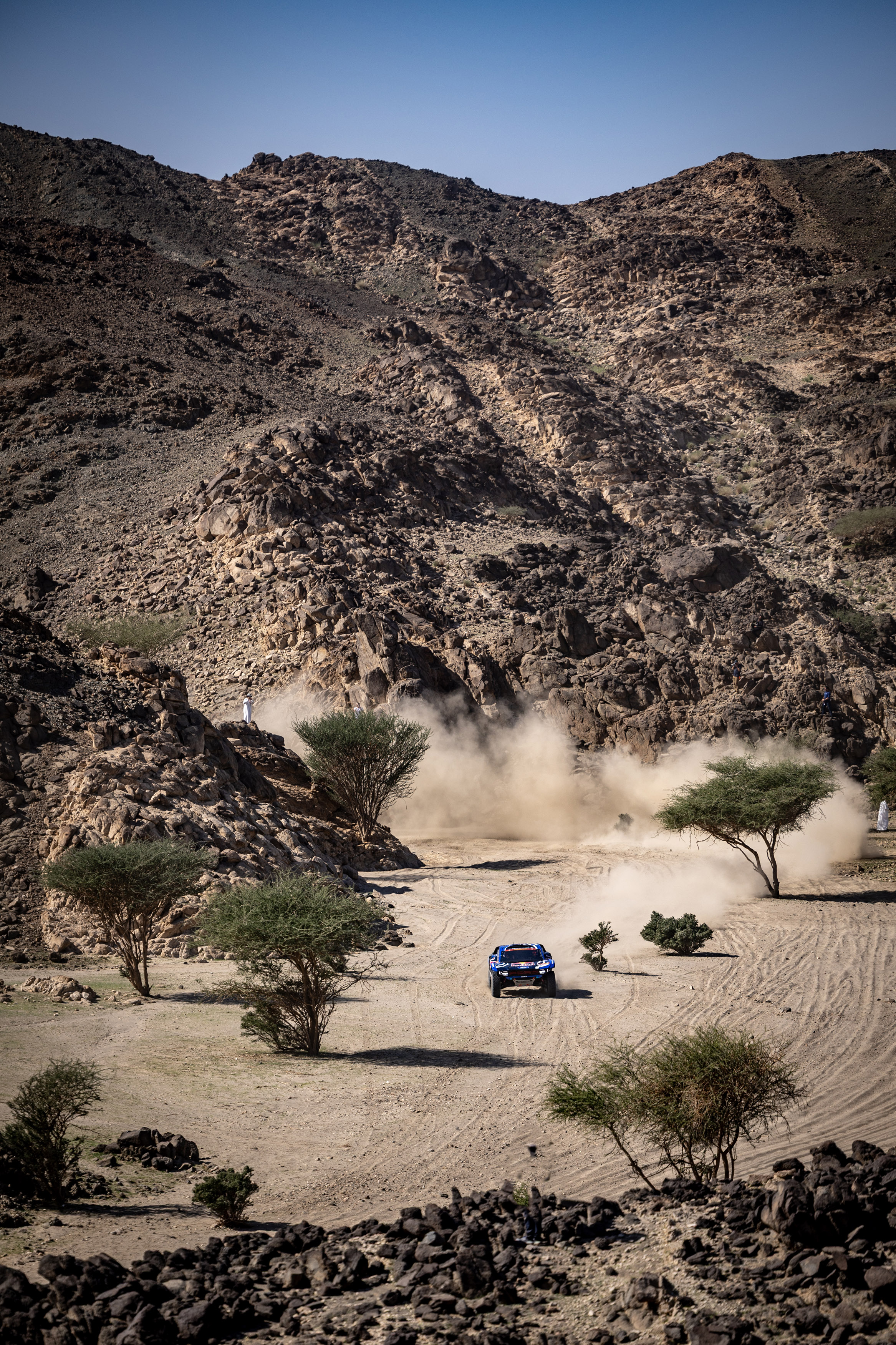 A Ford off-road vehicle drives through the desert. The backdrop of the photo includes a blue sky over a scenic ridge.