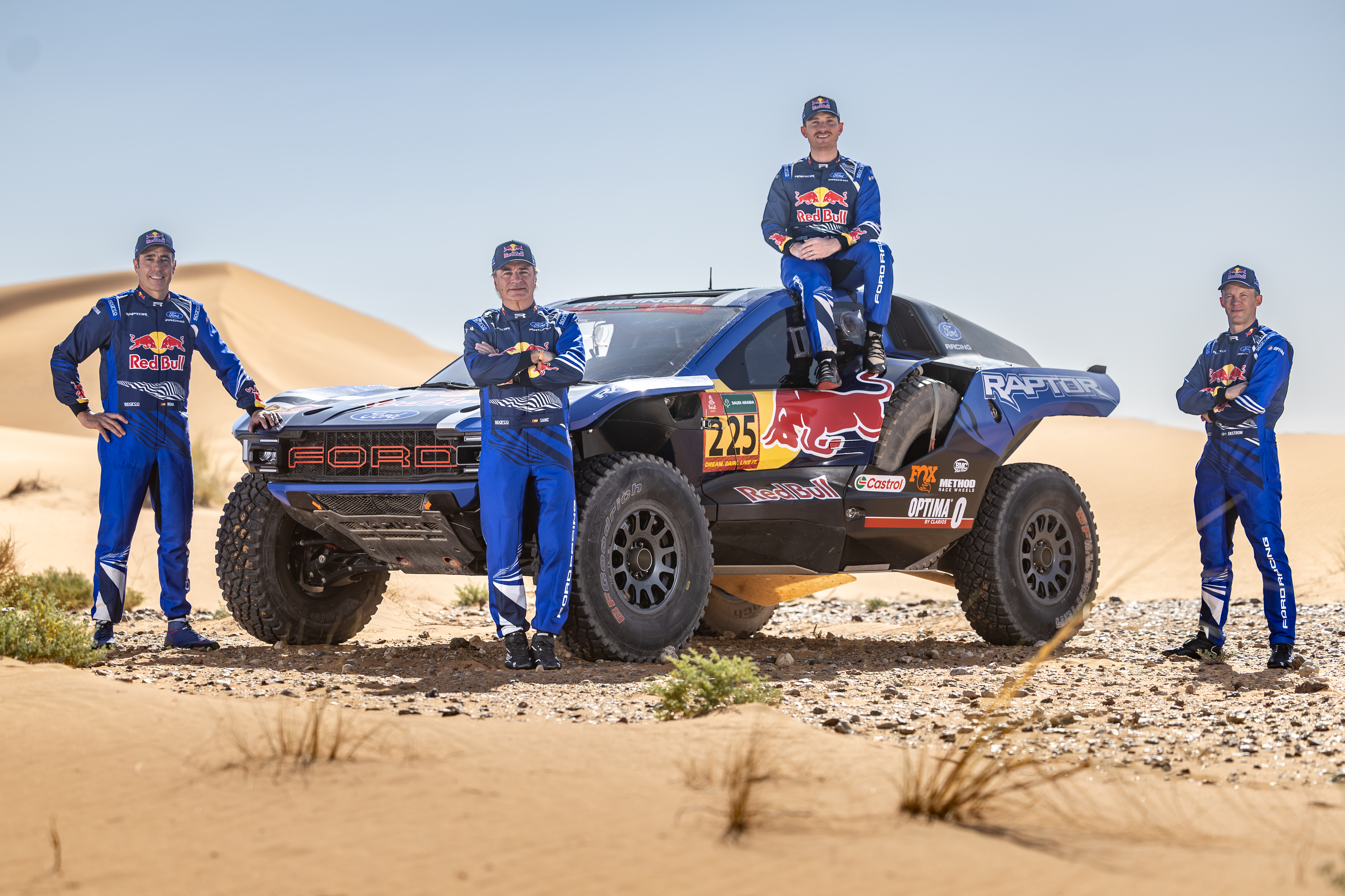 Four men in Ford Racing uniforms pose near an off-road Ford Racing vehicle in the desert.