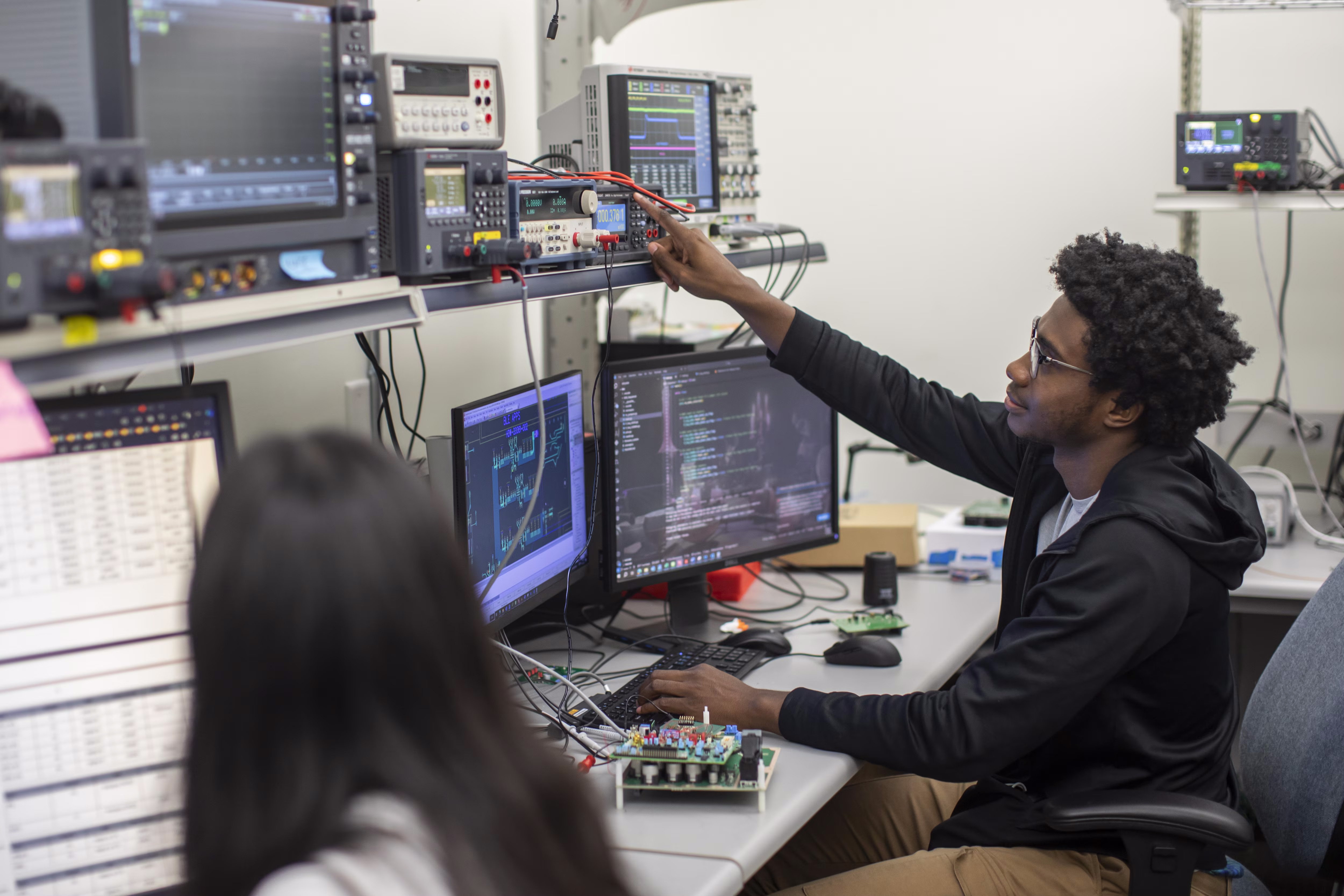 Two engineers sit at a desk with electronics upon it, including several computer monitors.